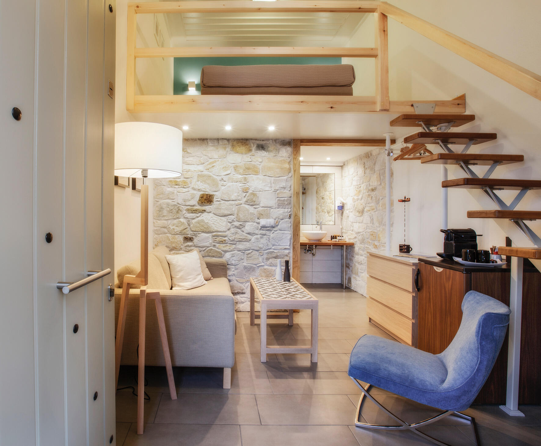 Μοντέρνο δωμάτιο boutique ξενοδοχείου με καθιστικό, ξύλινη σοφίτα, πέτρινους τοίχους και σύγχρονη επίπλωση σε γήινους τόνους. Modern boutique hotel room with a cozy sitting area, wooden loft, stone walls, and minimalist furnishings in natural tones.