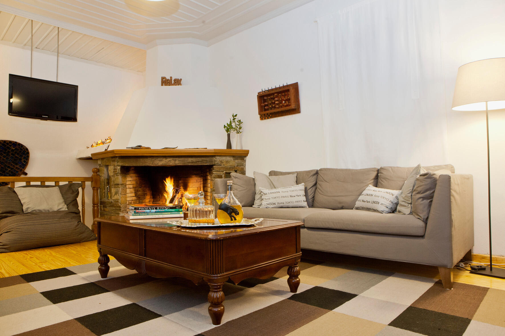 Καθιστικό με γκρι καναπέ, αναμμένο τζάκι από πέτρα και ξύλο, ξύλινο τραπεζάκι με βιβλία και ποτά, σε ζεστή και οικεία ατμόσφαιρα. Living room with grey sofa, lit stone-and-wood fireplace, wooden coffee table with books and drinks, creating a warm and invit