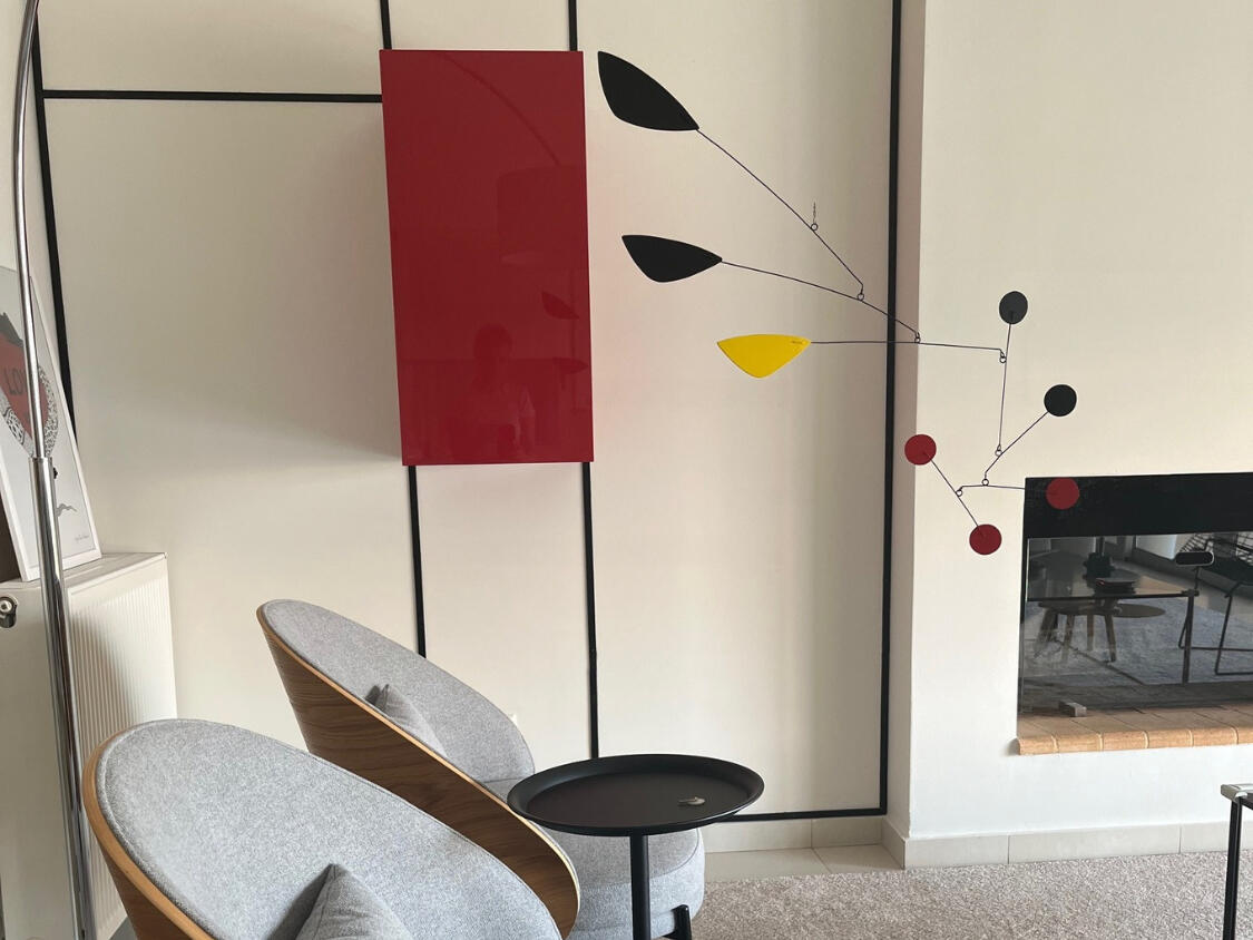 Mobile εμπνευσμένο από τον Alexander Calder, κρεμασμένο στο σαλόνι, με αφηρημένα γεωμετρικά σχήματα που αιωρούνται απαλά. Alexander Calder-inspired mobile hanging in a living room, featuring abstract geometric shapes gently suspended.